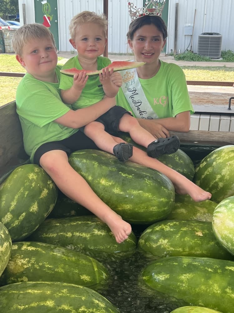 Two small children sit on a pile of watermelons, and an older child wearing a crown smiles behind them.