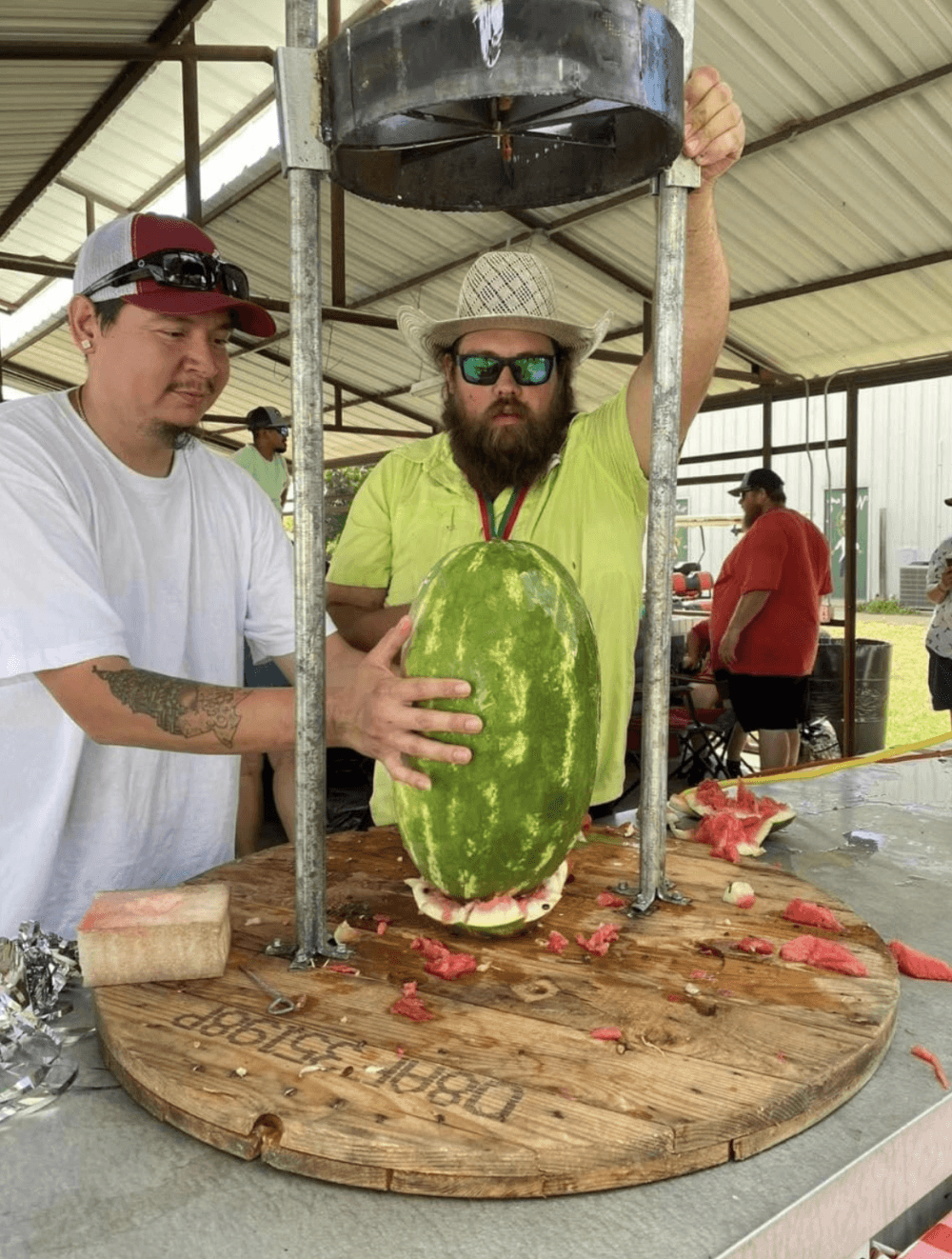 Two people set up a watermelon to be sliced with a large industrial slicer.