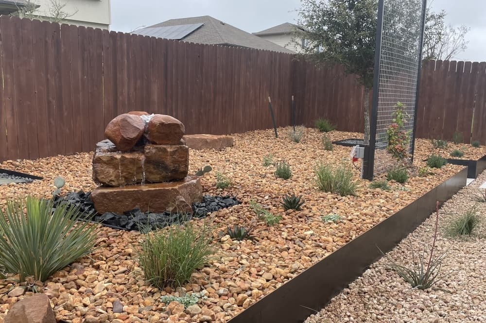 A backyard features a water feature with boulders and gravel, with agave plants nestled between the pebbles.  