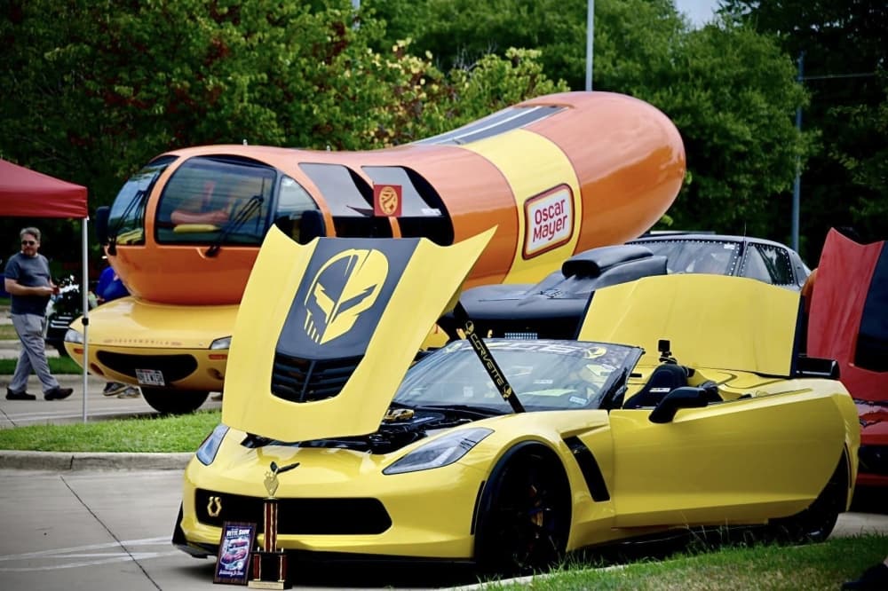 A yellow Corvette is shown in the foreground, and the Oscar Meyer Weinermobile is shown in the background.