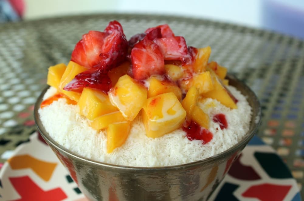 Bowl of ice cream substance topped with strawberries, mango and sauce. 