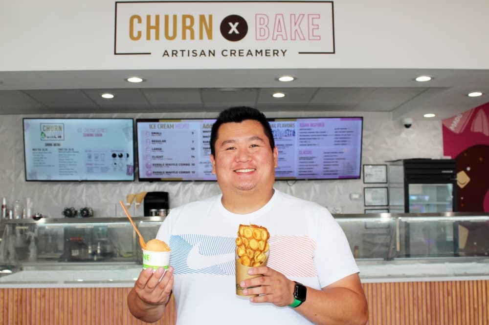 Male holding a waffle cone and a scoop of ice cream in a cup standing in front of a wall with Churn and Bake signage. 