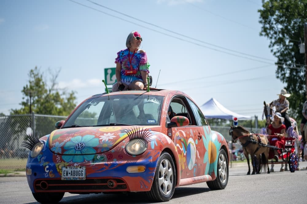A Volkswagon Beetle is shown painted in flowers with a woman on top of the car and people behind observing.