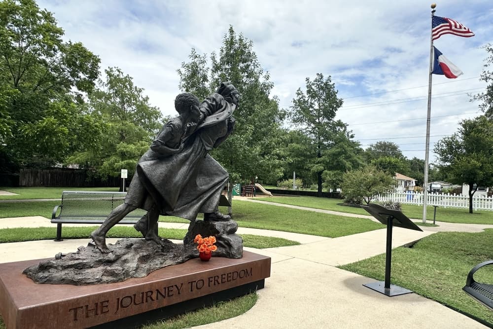 The statue is shown from the side on the left of the frame, with the Texas and American flags in the background on the right
