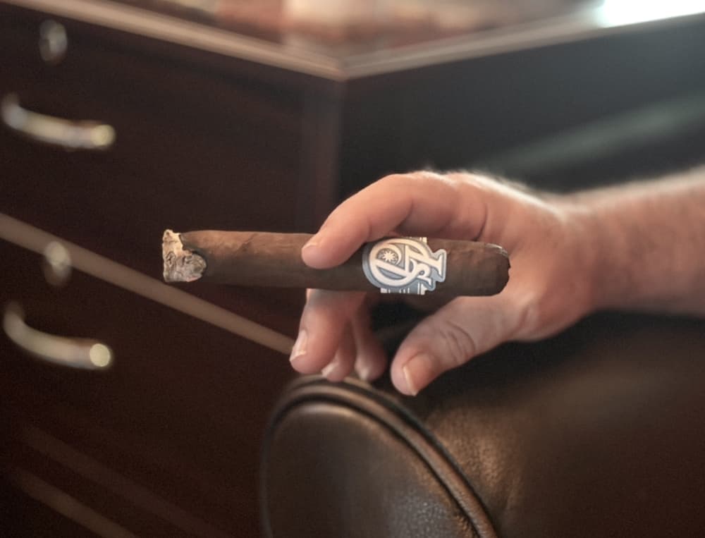 A close-up of a person's hand holding a cigar with the end-table humidor in the background.