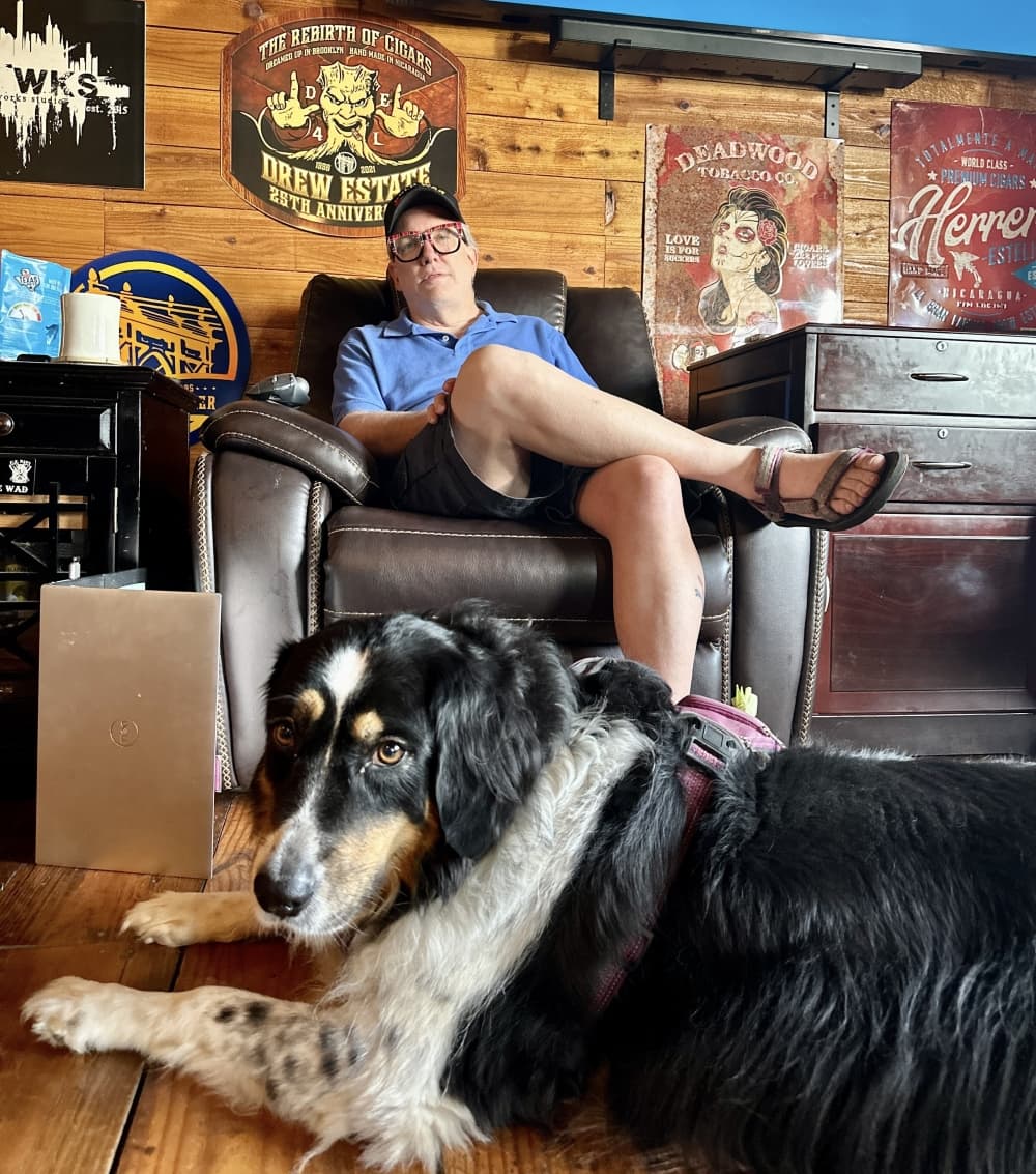 Rex Drake is shown in the background sitting in a leather chair, with his black and white dog, Mollie, at his feet in the foreground.