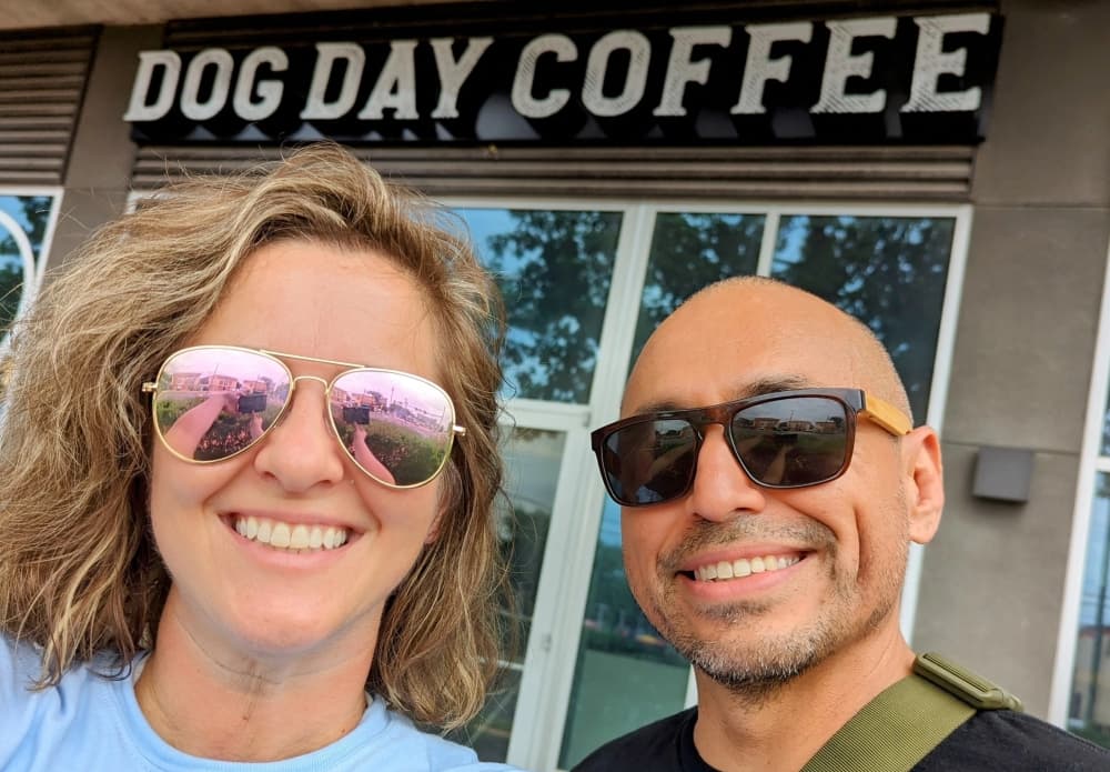 A man and woman wearing sunglasses taking a selfie outside of a coffee shop. 