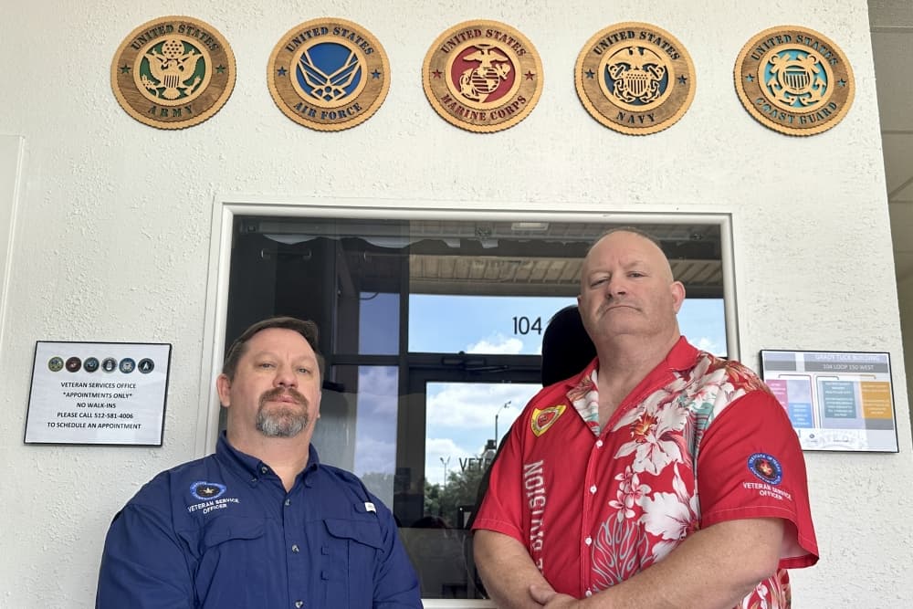 The two men stand side by side and pose for a photo in front of the entryway to the office, with military symbols above them.