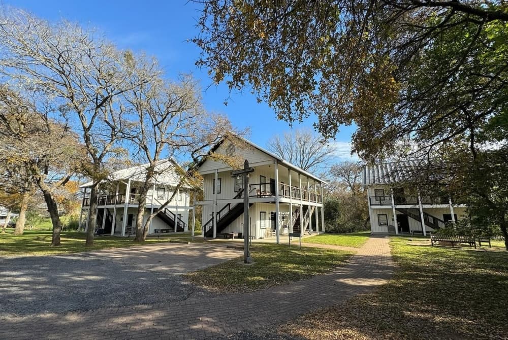 Two-story white buildings are shown amongst the trees with a blue sky.
