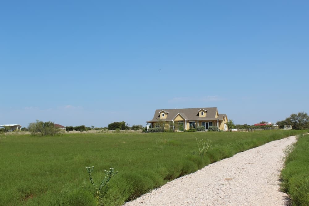 A gravel road brings drivers to a large yellow house with lots of green grass. 