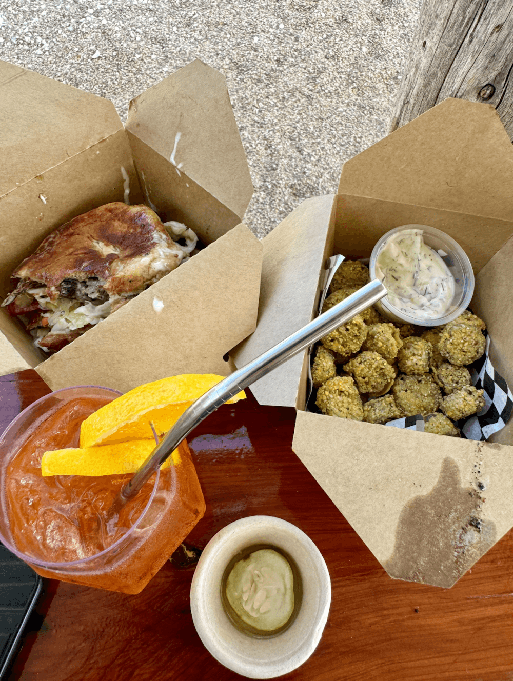 Two boxes are shown with the sandwich in one, okra in the other, next to a pink drink garnished with orange slices and a cup of aioli.