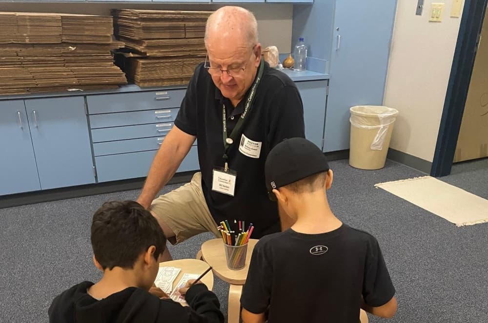 An older man kneels down next to two young students working on an assignment in school. 