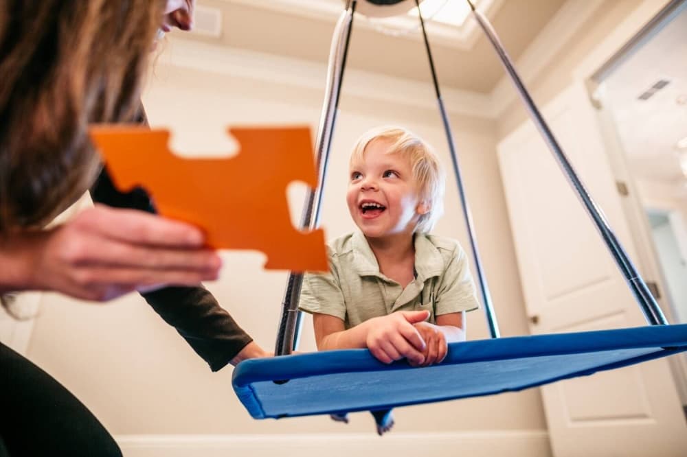 Young child swinging on a flat swing while an adult holds a puzzle piece. 
