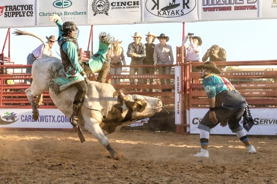 Picture of a cowboy on a bull in a rodeo as a clown stands nearby