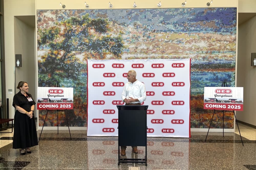 Mayor of Georgetown stands in front of a podium with an HEB backdrop while speaking to an official nearby. 