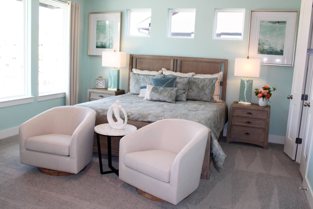 Bedroom with light sage green walls and bedspread with two off-white chairs. 