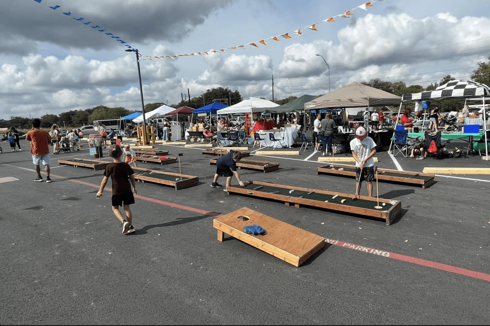 People are shown playing cornhole in the foreground with vendors in the background.