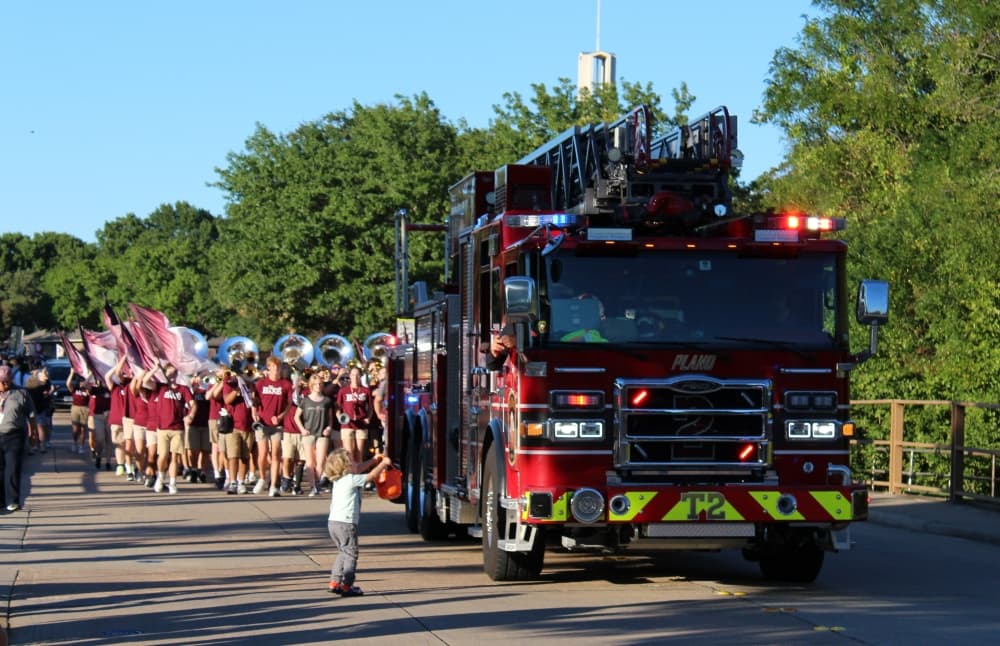 Fire truck leading a parade with a small child next to it holding a trick or treat bucket. 