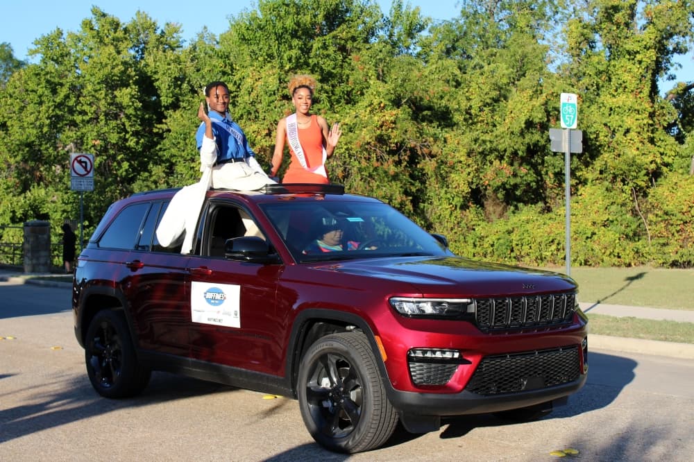 Teens sitting in sunroof of a car wearing homecoming king and queen sashes. 