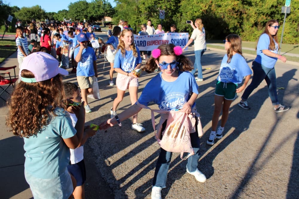 Children marching in a parade handing candy to parade goers. 