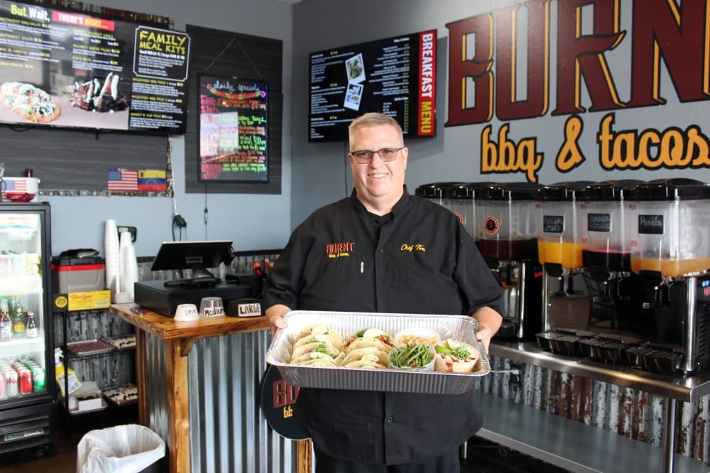 Man holding a tray of tacos and side dishes in a restaurant with bbq and tacos sign behind him. 