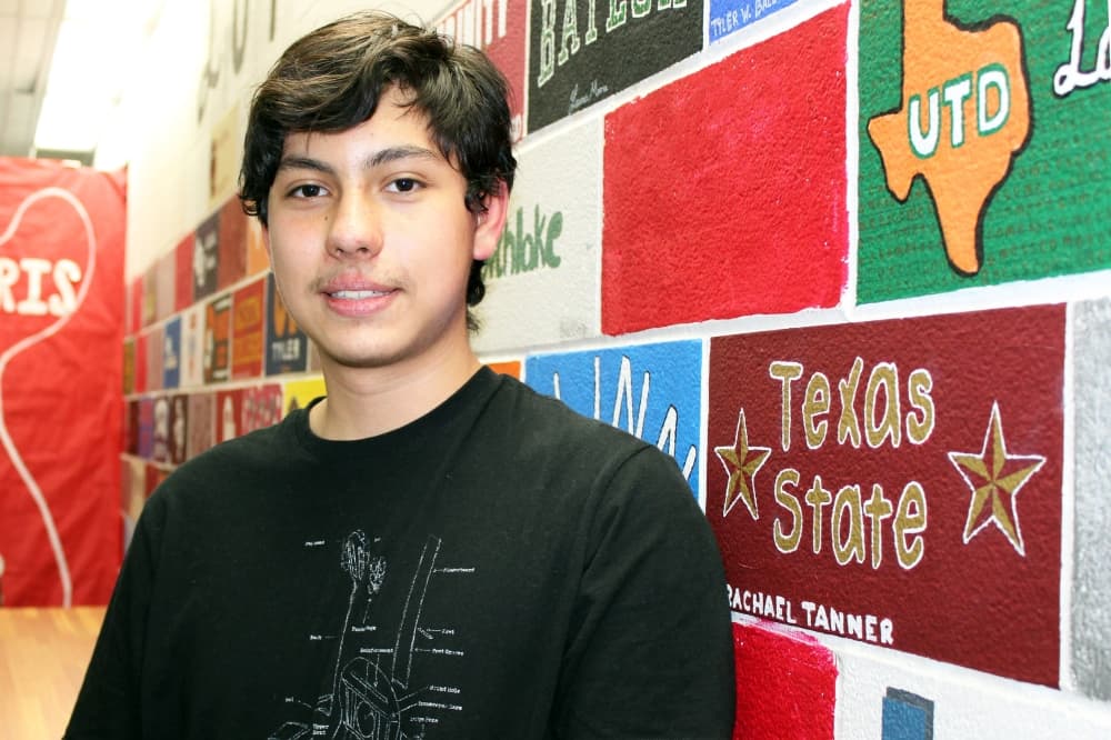Male standing in front of a wall with college logos painted on it. 