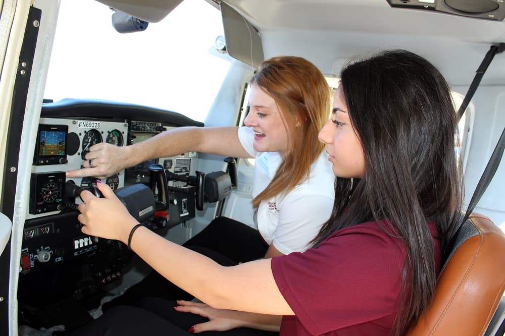 Female wearing white shirt pointing at plane instrument panel while girl wearing maroon shirt looks on. 