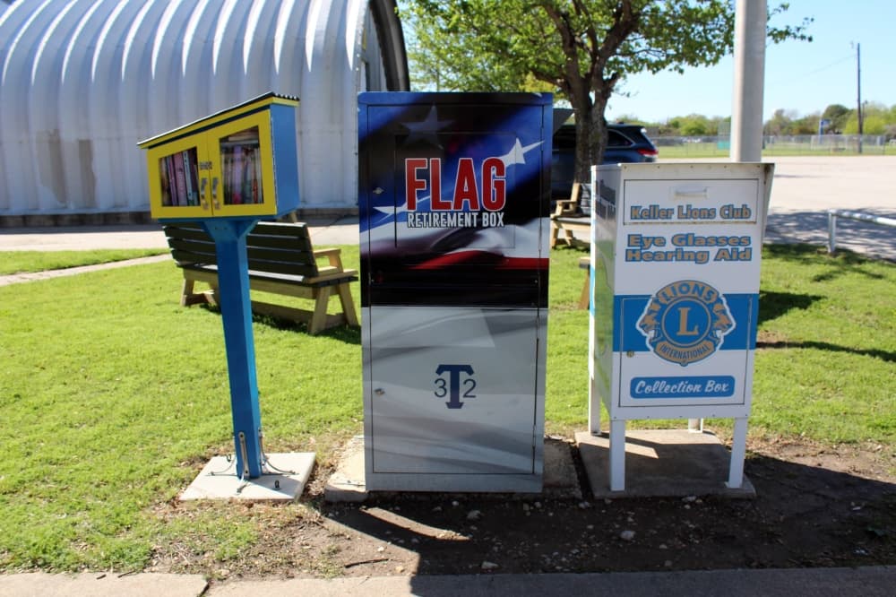Little library on a pole, flag donation receptacle and eyeglass donation receptacle side by side outside a metal building.  