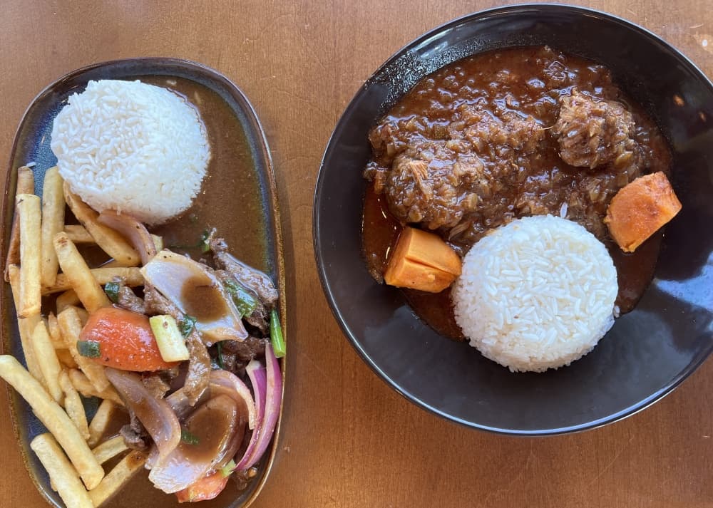 On the left, a dish called lomo saltado sits with a mound of white rice next to French fries, orange sweet potatoes, a slices of steak coated in a dark brown sauce. On the right is another dish called adobo limeño. The second plate features a mound of rice, orange sweet potatoes and pork in rounded balls with a dark brown sauce. 