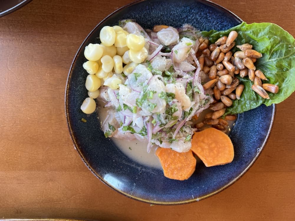 A blue bowl holds a ceviche dish made of pale fish covered in lime juice. The fish sit on a green piece of lettuce with brown seeds and two slices of sweet potatoes. On the side of the fish, pale white Peruvian corn is piled up.