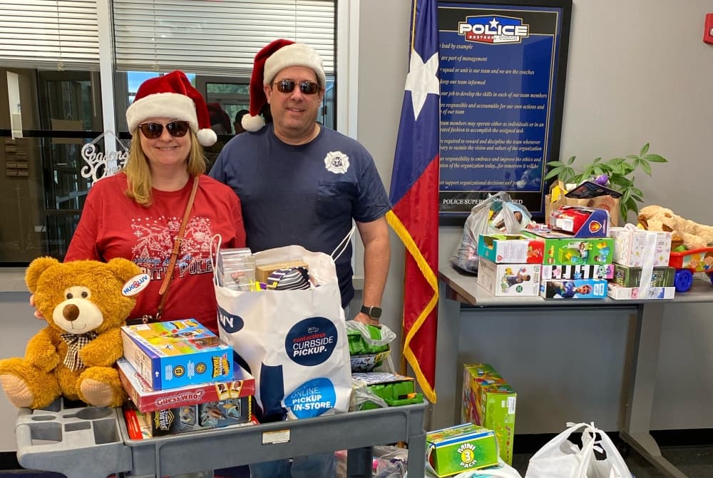 Two people wearing Santa hats pose in front of toys from a previous Operation Blue Santa.