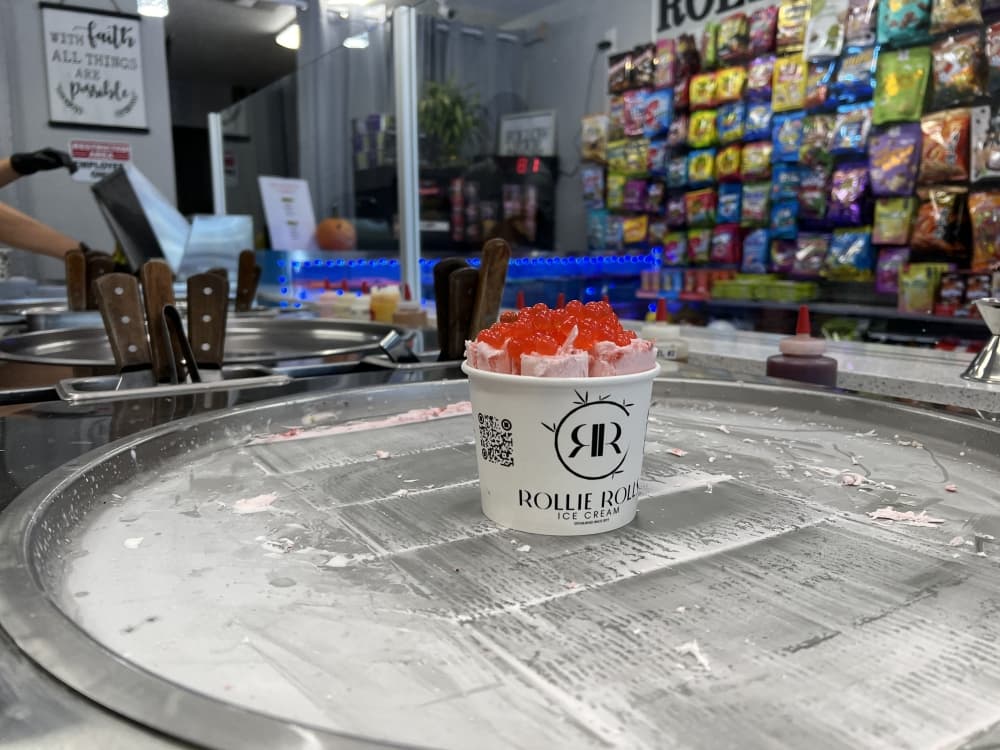 A cup of ice cream sits on top of a circular grey pan. Inside the cup, the ice cream is rolled into cylinder shapes and topped with bright red boba. Behind the cup, a wall of assorted ramen packages lines the store.