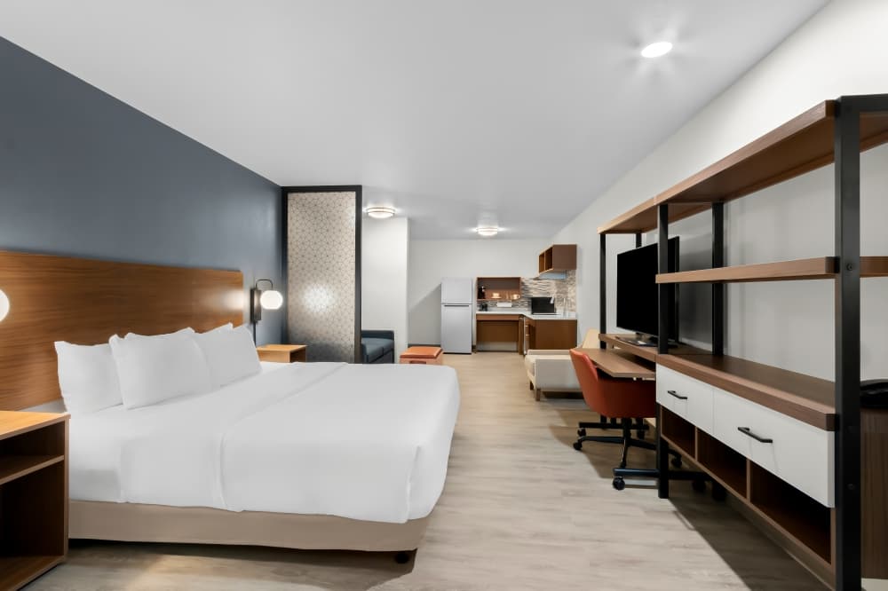 The inside of a room similar to what will be inside the hotel is shown with white bedding, a dark accent wall, shelving and a kitchenette in the background.