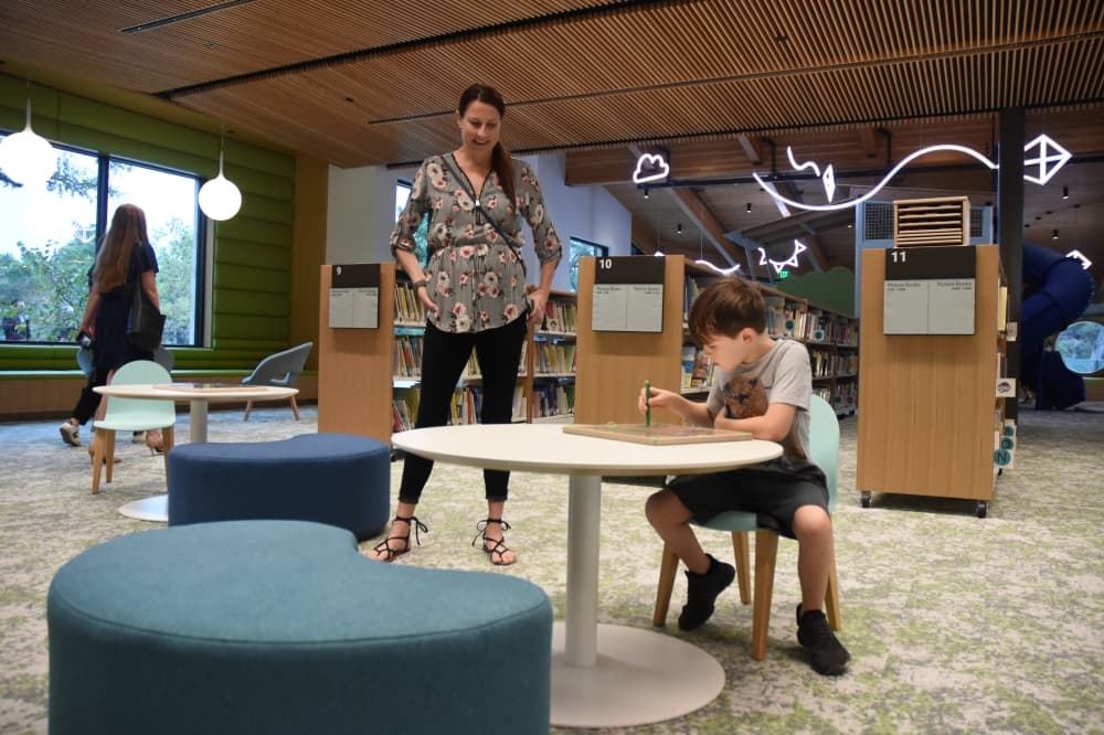 A woman stands to the side as her son sits at a table and plays with a game. 
