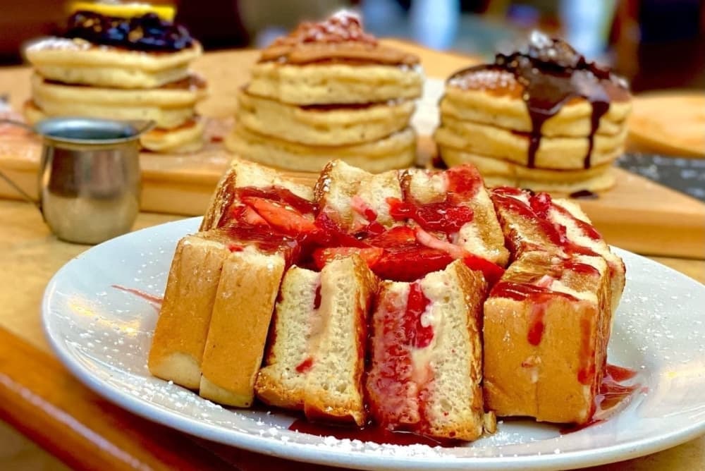 The French toast is cut into slices and covered in strawberries in the foreground, with a pancake flight in the background.