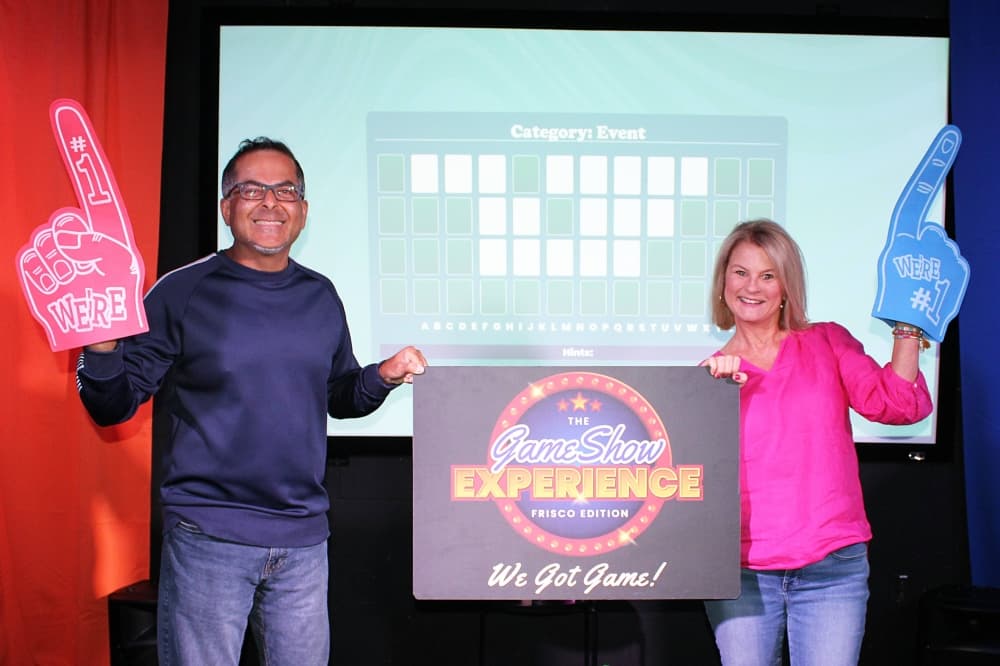 Male and female wearing foam fingers holding a The Game Show Experience sign between them. 