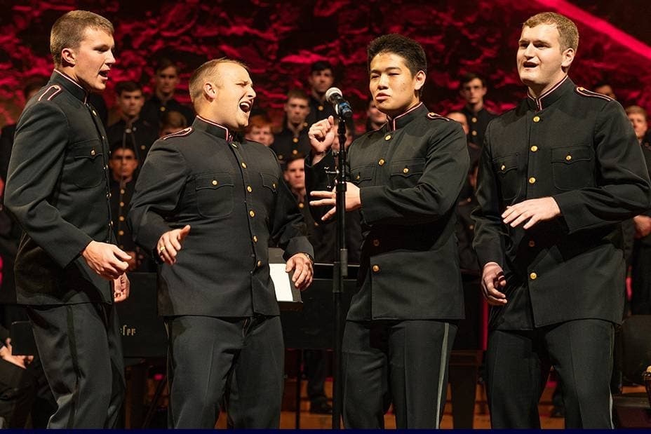 Four males wearing military style uniforms singing on stage. 