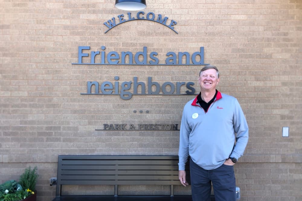 Male standing in front of a brick wall with 'Welcome Friends and Neighbors' signage. 