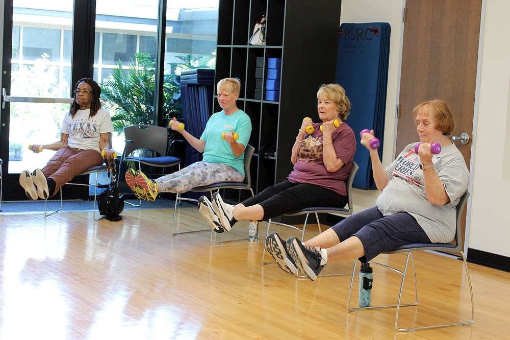 Four females sitting in chairs lifting legs and lifting weights with their arms. 