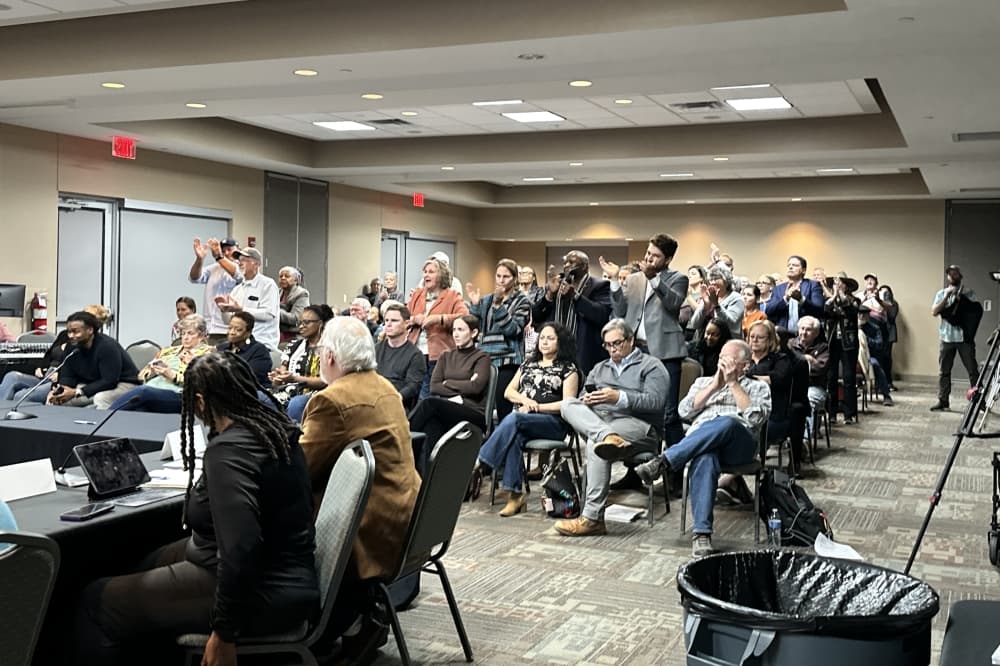 The crowd is shown in a packed room during the meeting.