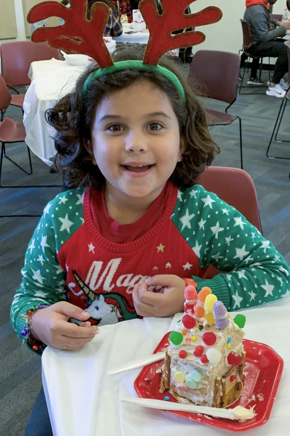 A child wearing a reindeer hat with antlers smiles in front of their decorated graham cracker house.