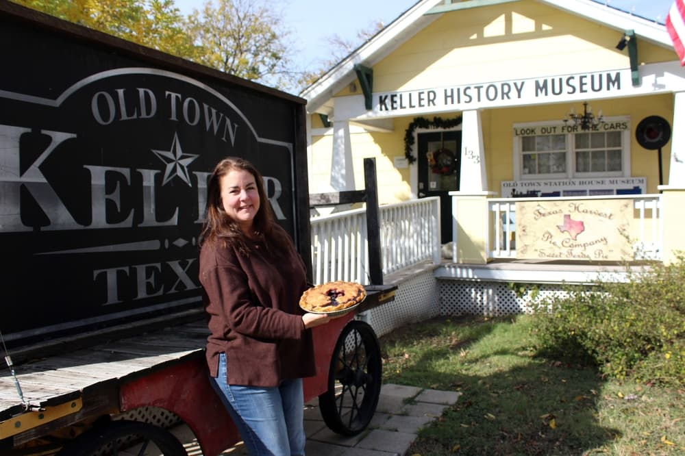 Female holding a pie while leaning against a sign in front of a house with Keller History Museum signage. 