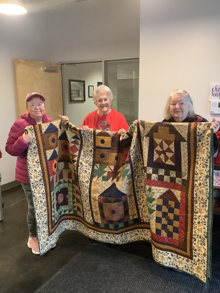 An image of three seniors holding up a quilt. 