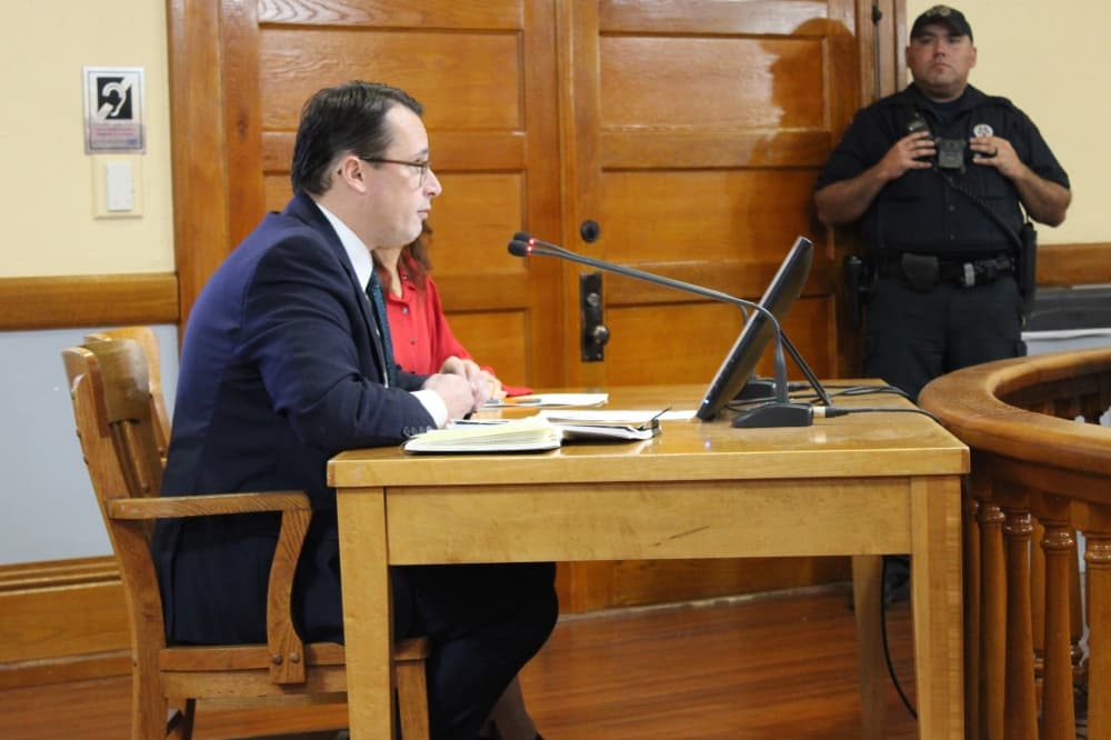 A man sits in a chair beside a woman in a courtroom, speaking into a microphone. 