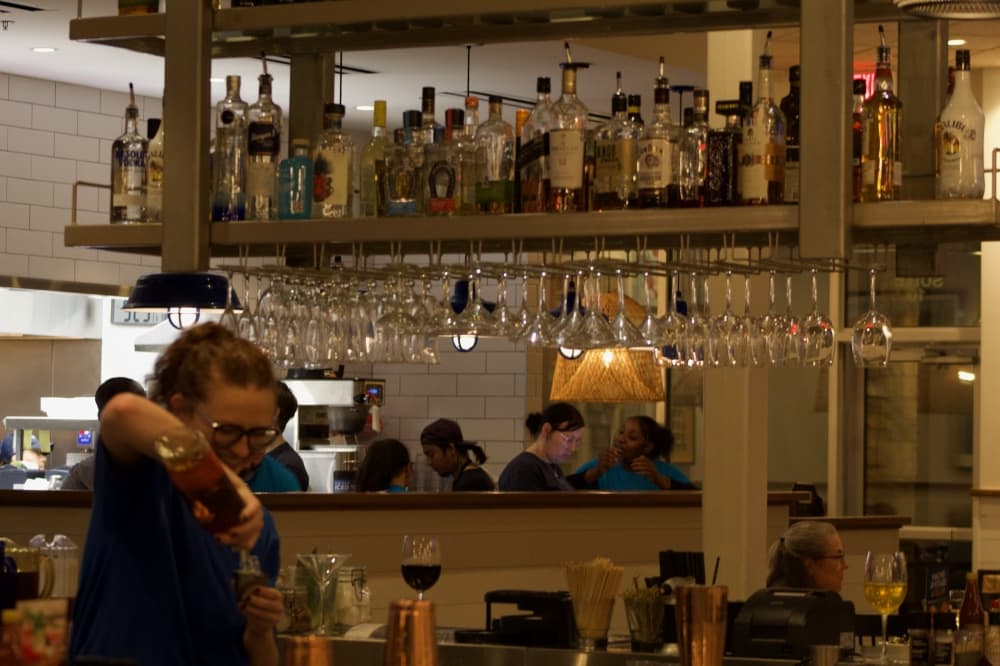 Girl making a drink at bar with beverages on shelf behind her.