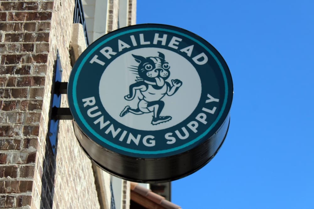 Circular plastic sign attached to a building featuring Boston Terrior wearing running attire with Trailhead Running Supply lettering. 