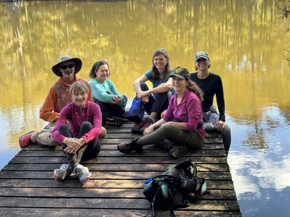 A group of people sit on a wooden dock with autumn trees reflected in the water