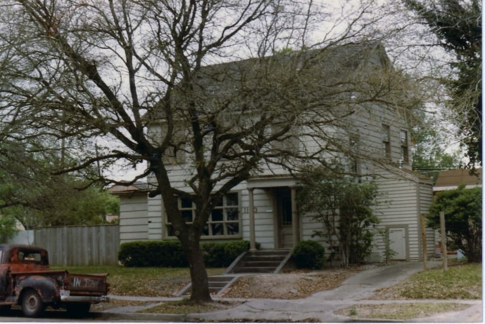 A white home with a tree in front of the home, a red truck parked on the curb of the home.