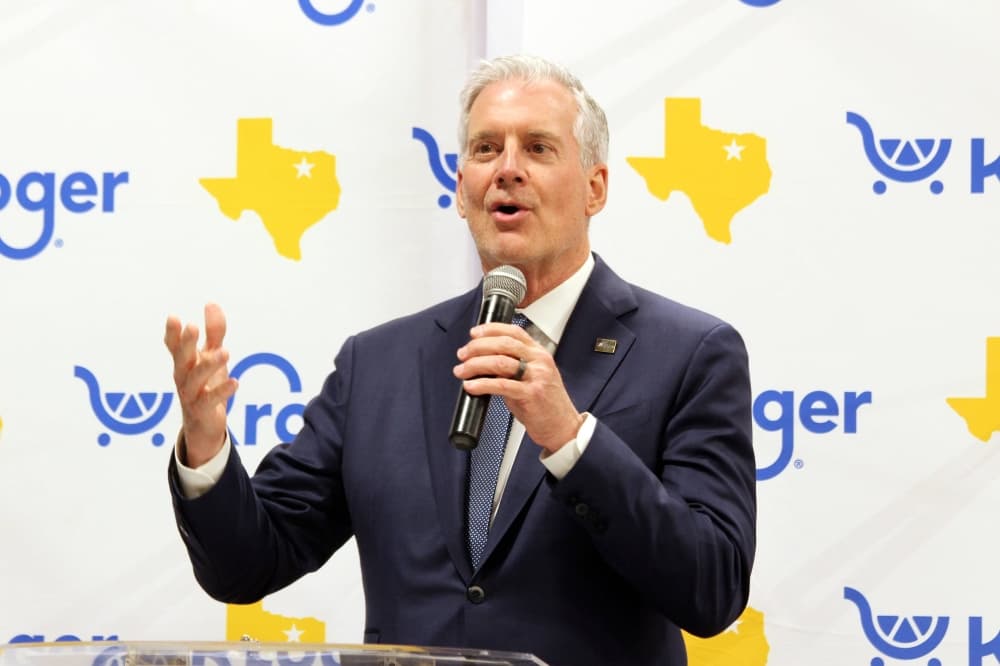 Male holding a microphone in front of a photo opp backdrop with Kroger logos. 