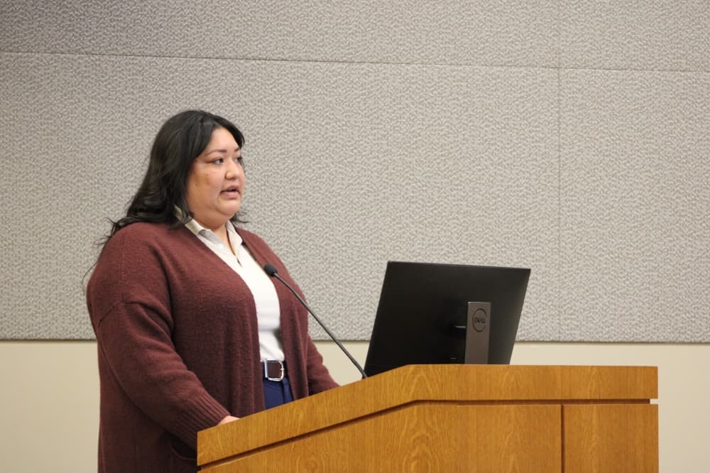 A woman speaks into a microphone at a podium during a council meeting. 